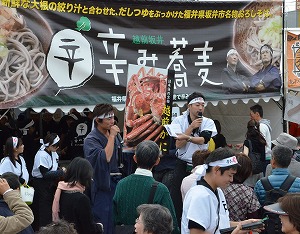 辛み蕎麦のブース前に並ぶ人たちにボードを掲げて越前がにをＰＲする後藤隊長ら＝９日、愛知県豊川市